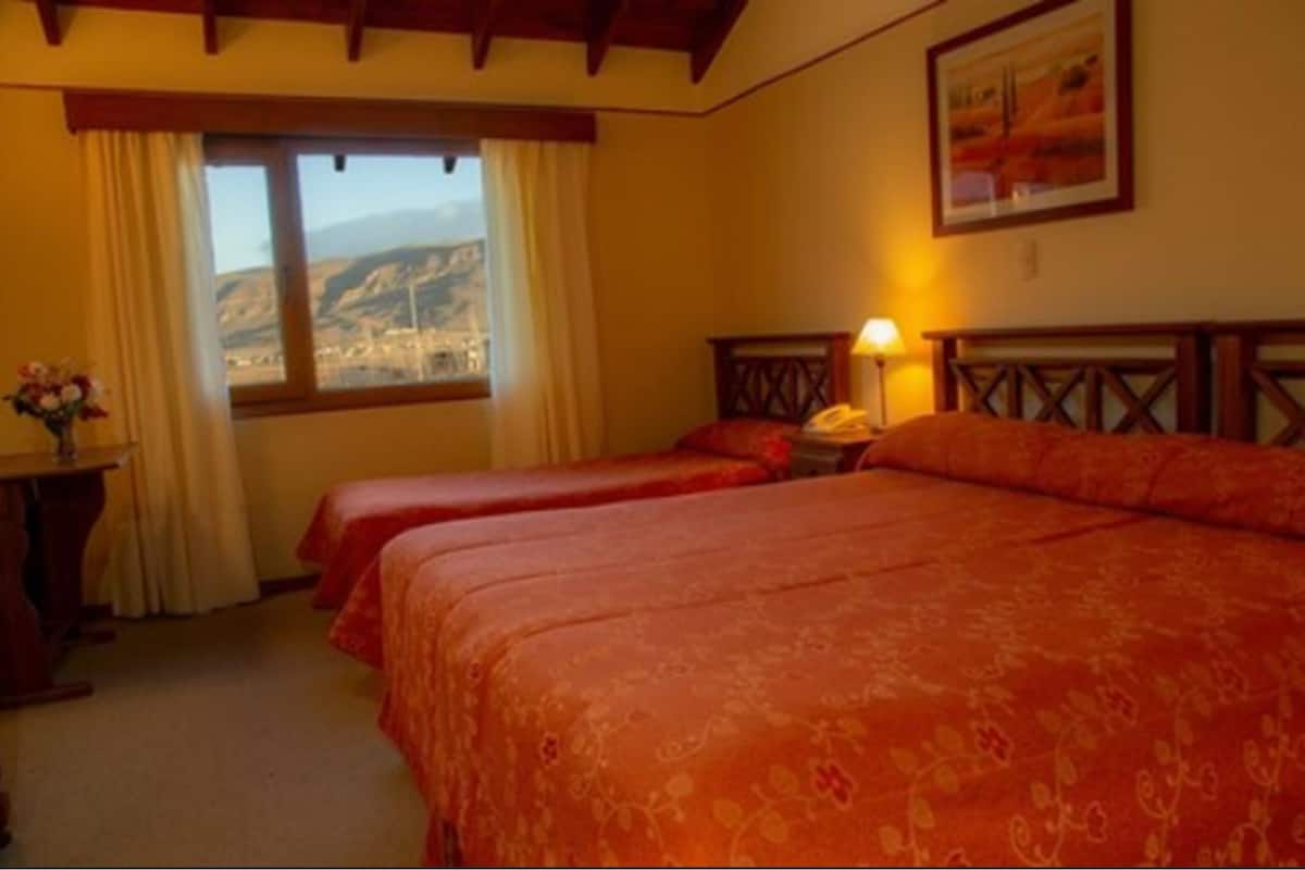 A hotel room with two beds, a window with a view, a bedside table with a lamp, and a painting on the wall.