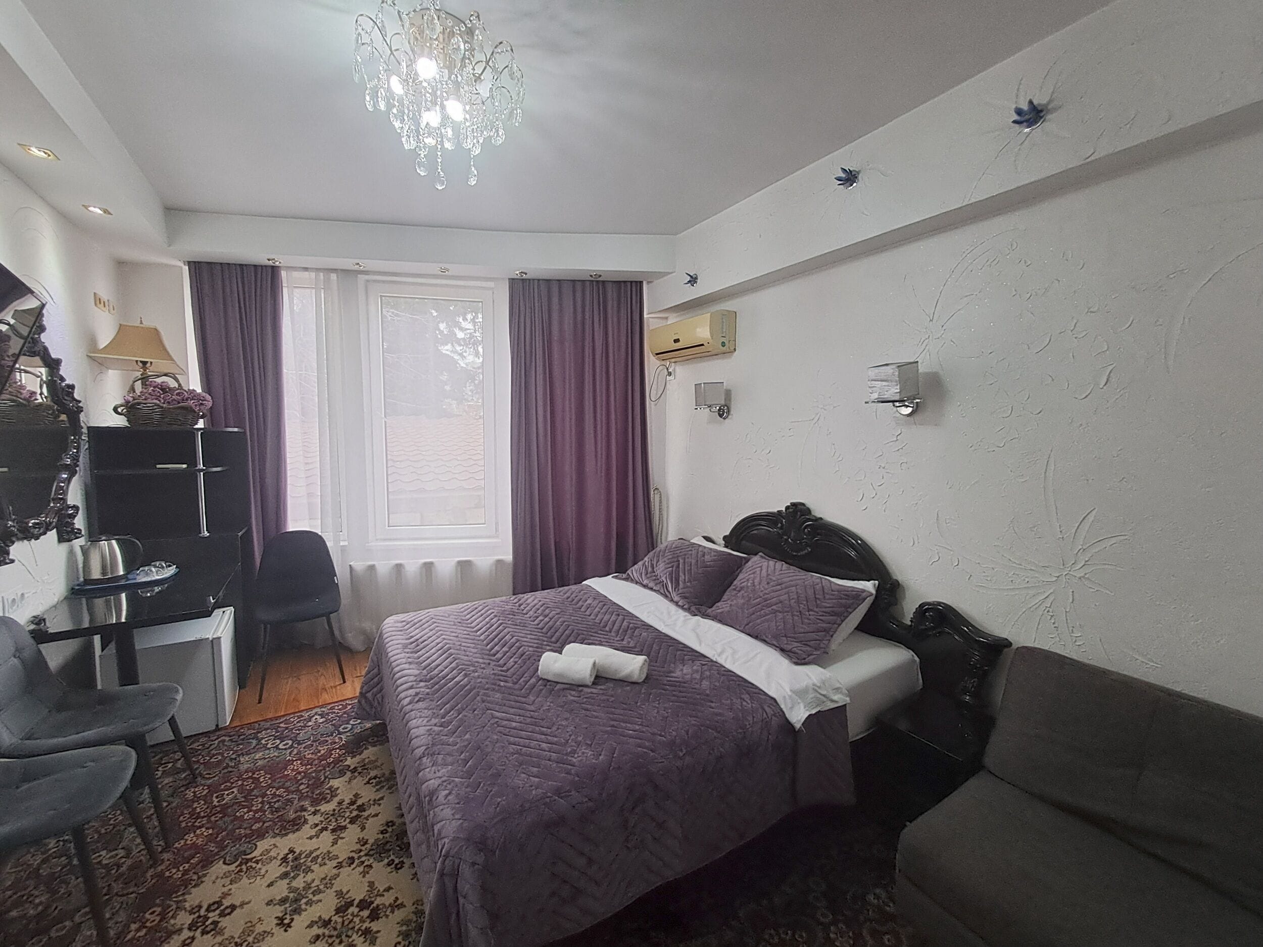 A hotel room with a bed, a desk, a chair, a sofa, a chandelier, and a window with curtains.