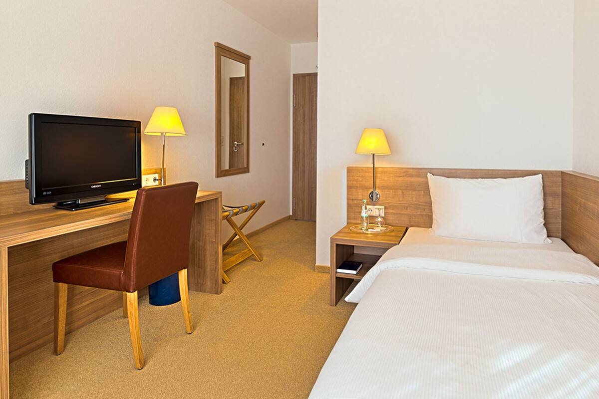 A hotel room with a bed, a desk with a computer, a chair, and two bedside lamps.