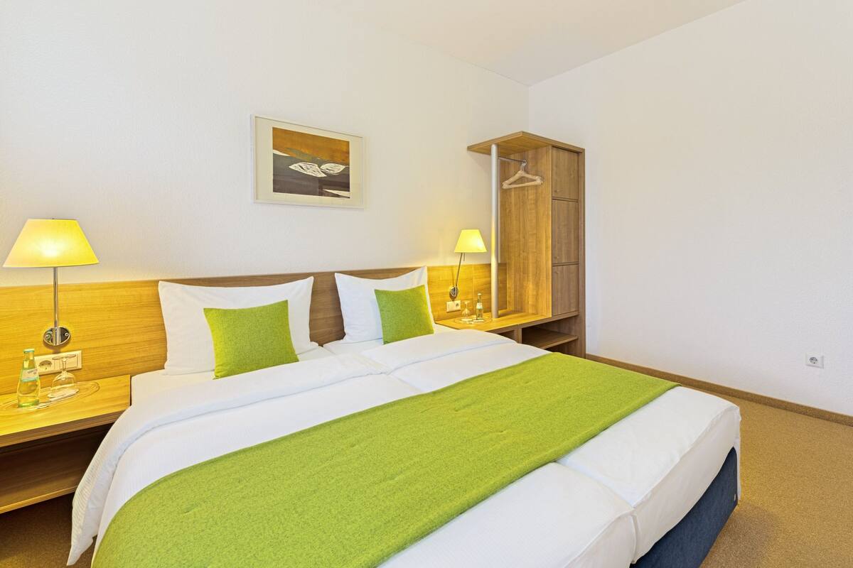 A hotel room with a bed, two bedside tables, a nightstand, a wooden wardrobe, a door, and a wall-mounted picture.