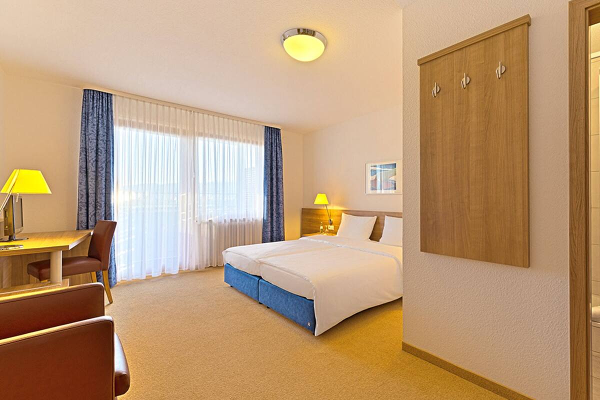 A hotel room with a bed, a desk with a chair, a desk lamp, a nightstand with a lamp, a wardrobe, and a bathroom with a sink and toilet.