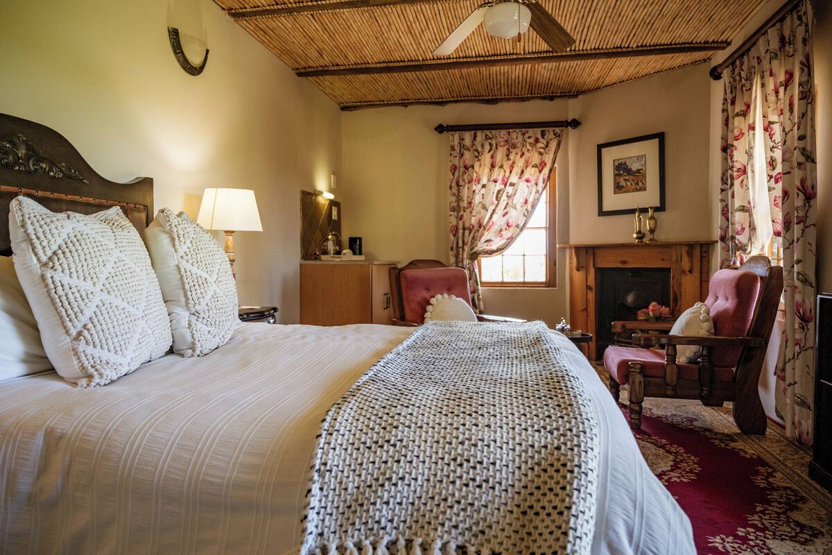 Un dormitorio con una cama grande, cabecera de madera, mesita de noche con lámpara, una silla, chimenea y ventana con cortinas florales.
