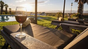 A glass of red wine on a stone table with a pool and lounge chairs in the background.
