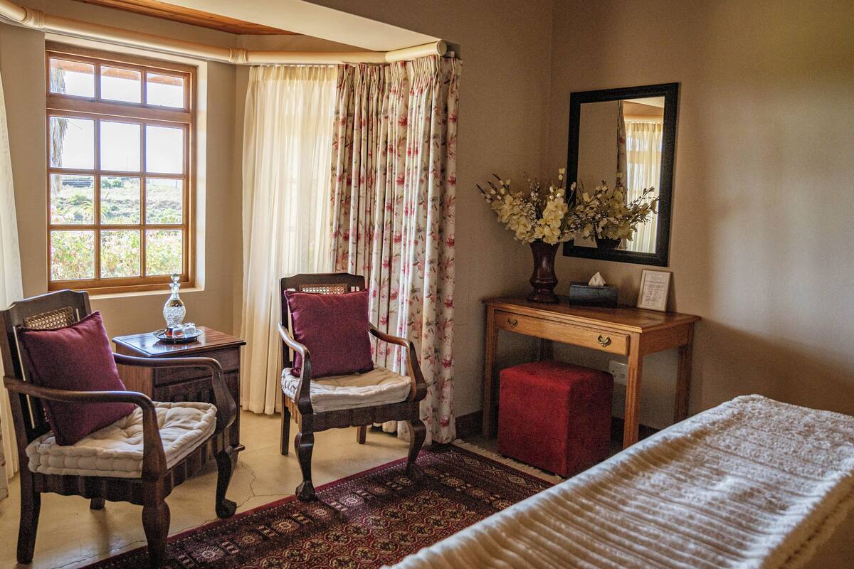 Una habitación con una cama, dos sillas, un escritorio, un espejo y una ventana con cortinas.