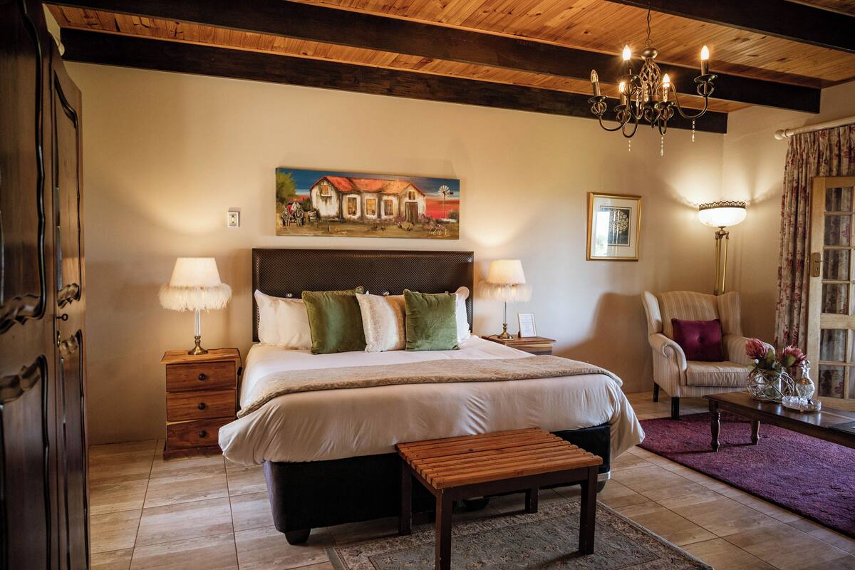 Un dormitorio con una cama grande, cabecera de madera, dos mesitas de noche con lámparas, un banco de madera, un cuadro en la pared y una silla con un jarrón de flores.