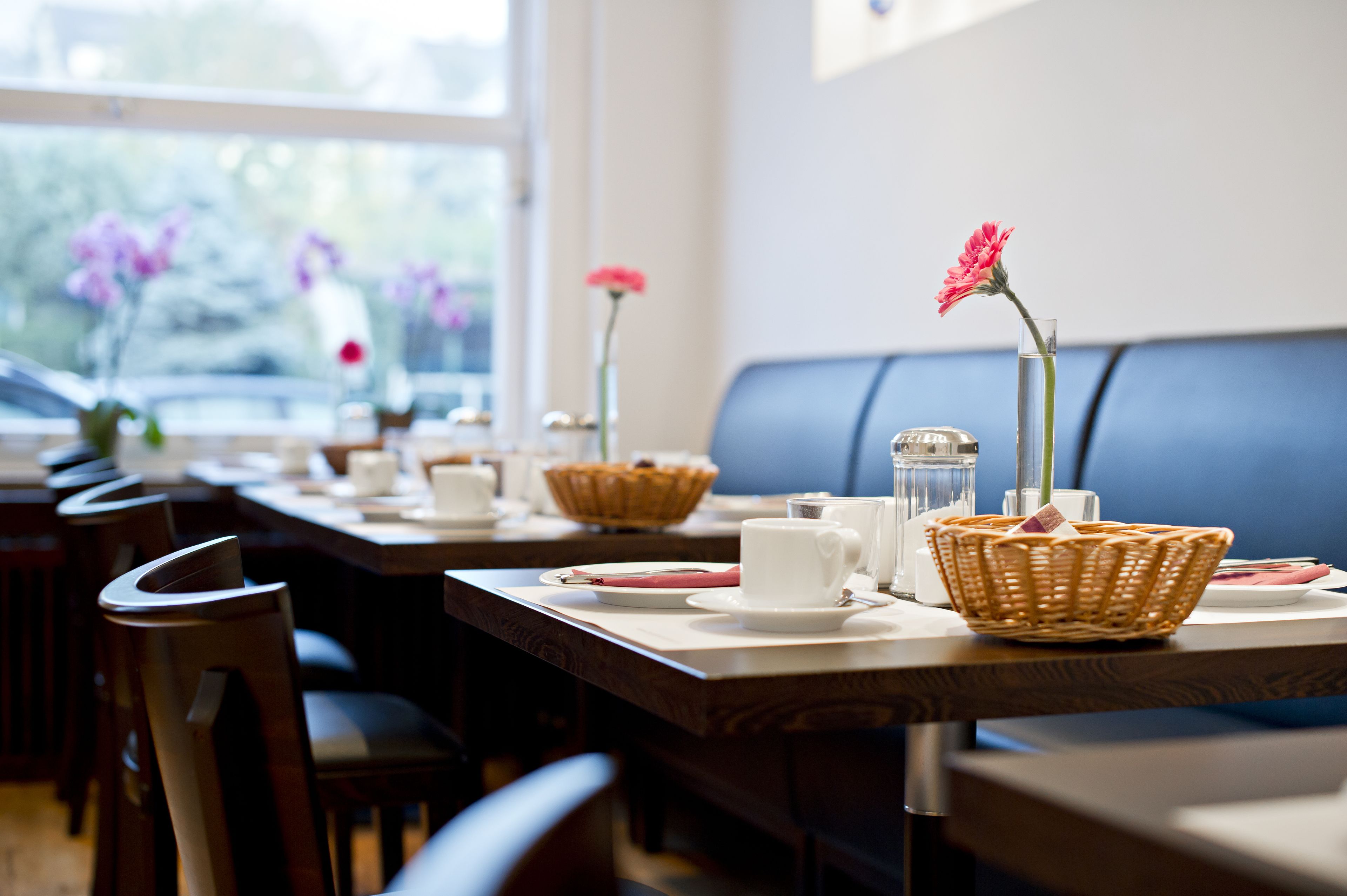 Un restaurante con mesas de madera, tazas blancas y cestas de mimbre con flores.