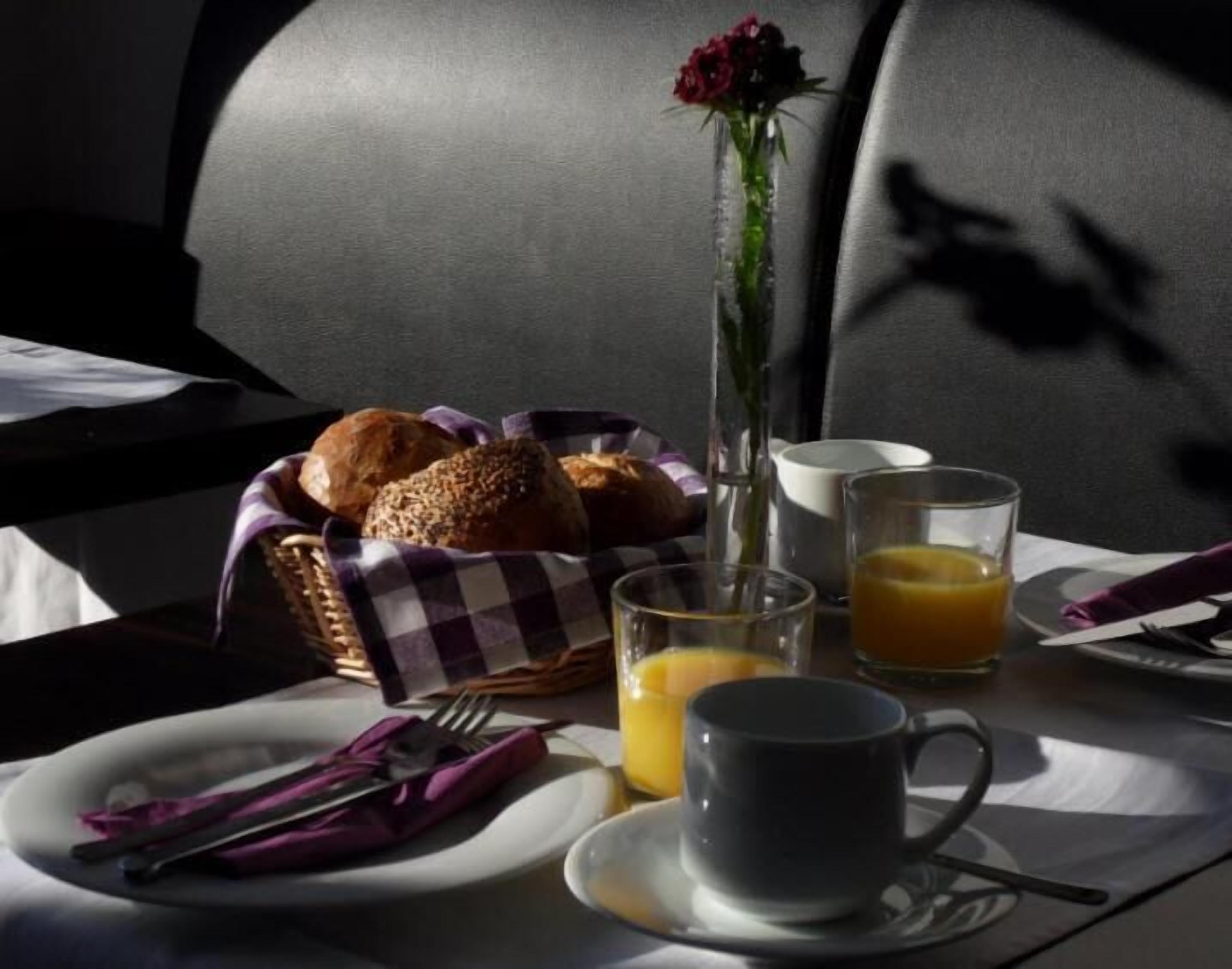 A breakfast setting with bread, a vase with a flower, a glass of orange juice, and a cup on a tray.