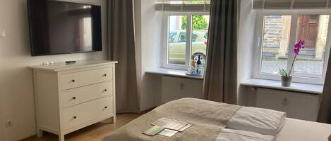 Un dormitorio con cama, televisor en una cómoda y ventanas con cortinas.