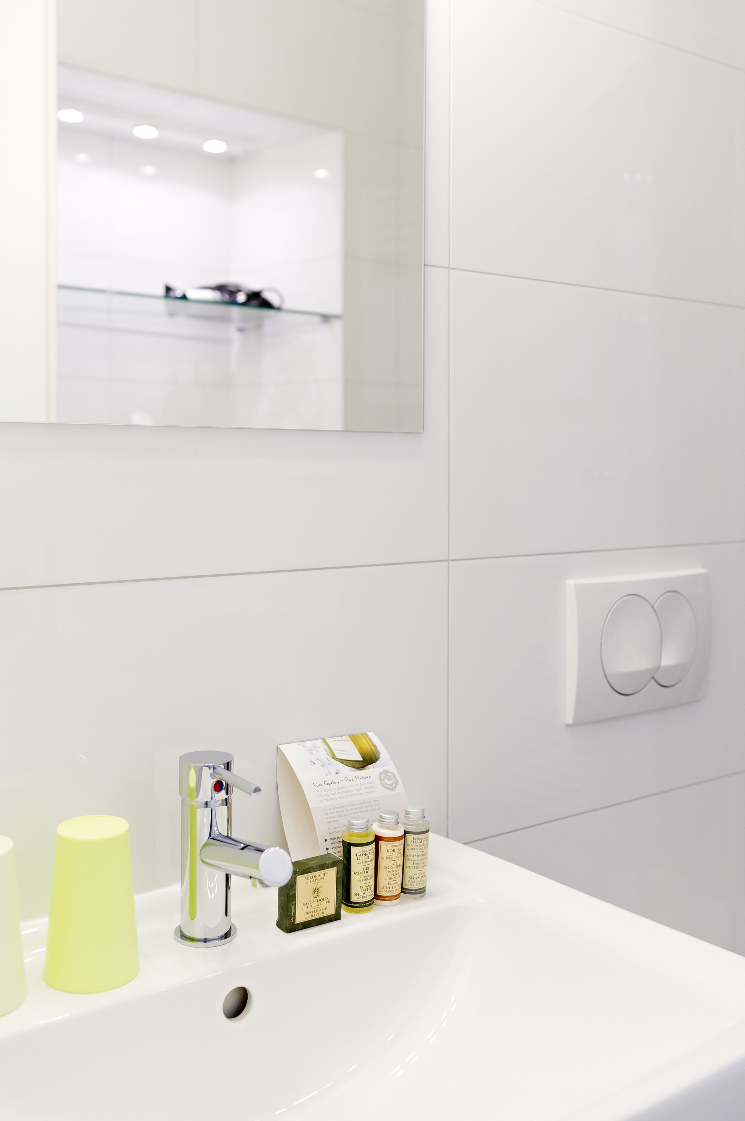 A modern bathroom with a white sink, a faucet, and a mirror. There are toiletries on the sink and a light switch on the wall.