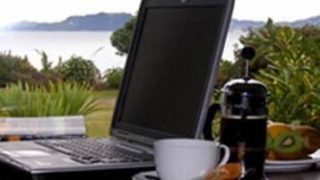 A laptop on a table with a cup and a beverage container, set against a backdrop of greenery and a body of water.