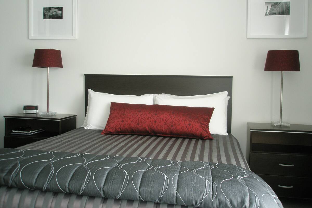 A neatly made bed with a striped bedspread and two pillows, one red and one white, flanked by two bedside tables with lamps.