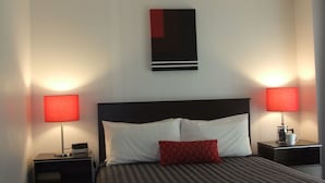 A hotel room with a bed, two bedside tables, a red lamp on each table, a red and black abstract painting on the wall, and a white pillow on the bed. - 83 By the Sea Motor Lodge  (Lower Hutt)
