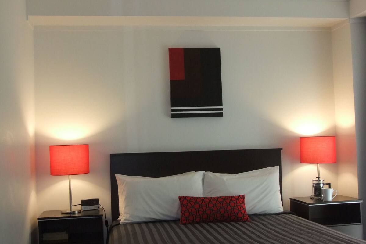 A hotel room with a bed, two bedside tables, a red lamp on each table, a red and black abstract painting on the wall, and a white pillow on the bed.