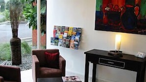A living room with a brown leather sofa, a black coffee table with a magazine, a black side table with a lamp, a wall-mounted art piece, and a bulletin board with various items pinned to it. - 83 By the Sea Motor Lodge  (Lower Hutt)