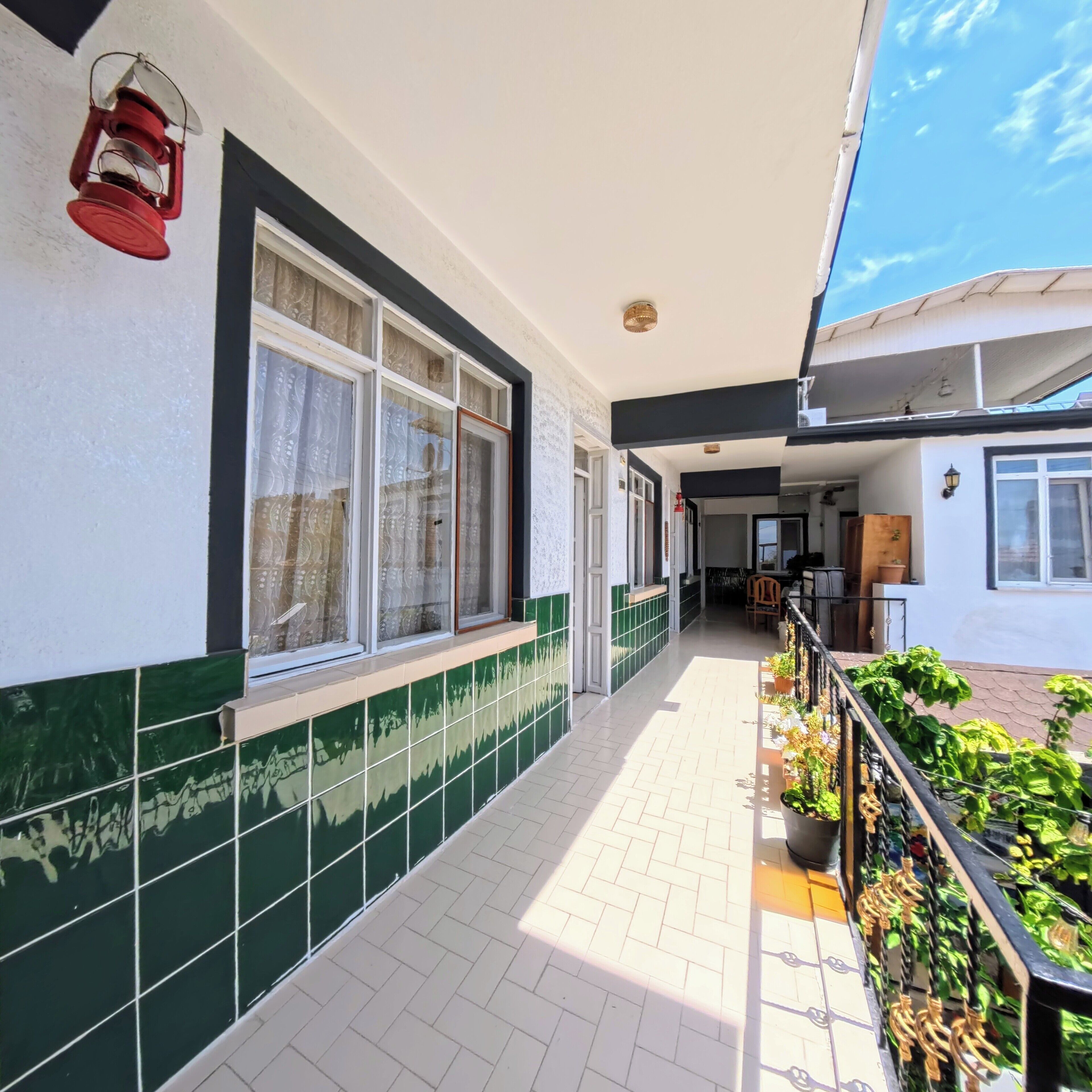 A corridor with tiled walls, large windows with curtains, and a balcony with potted plants.