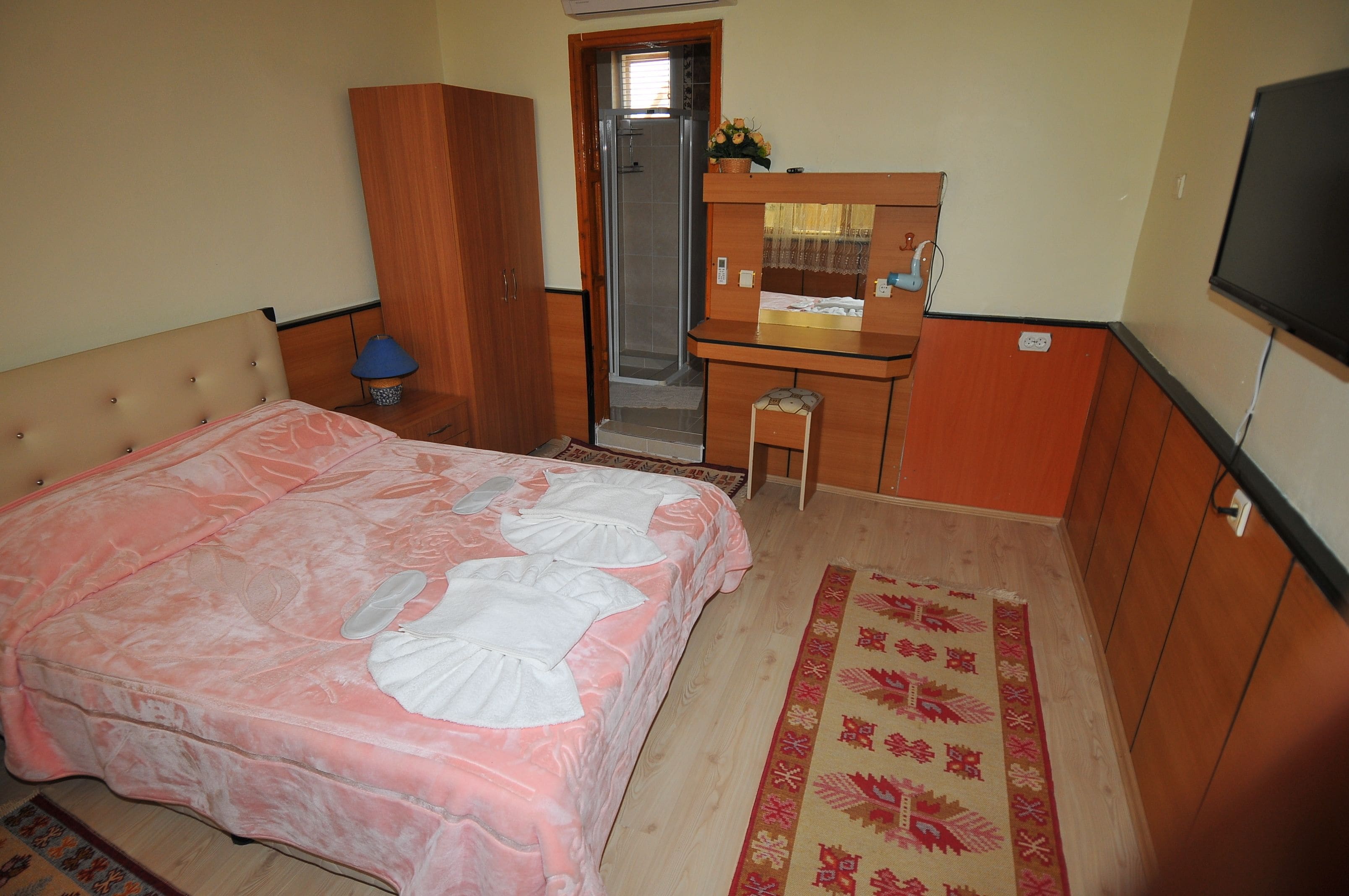 A bedroom with a bed, a wooden wardrobe, a desk with a mirror, and a television.