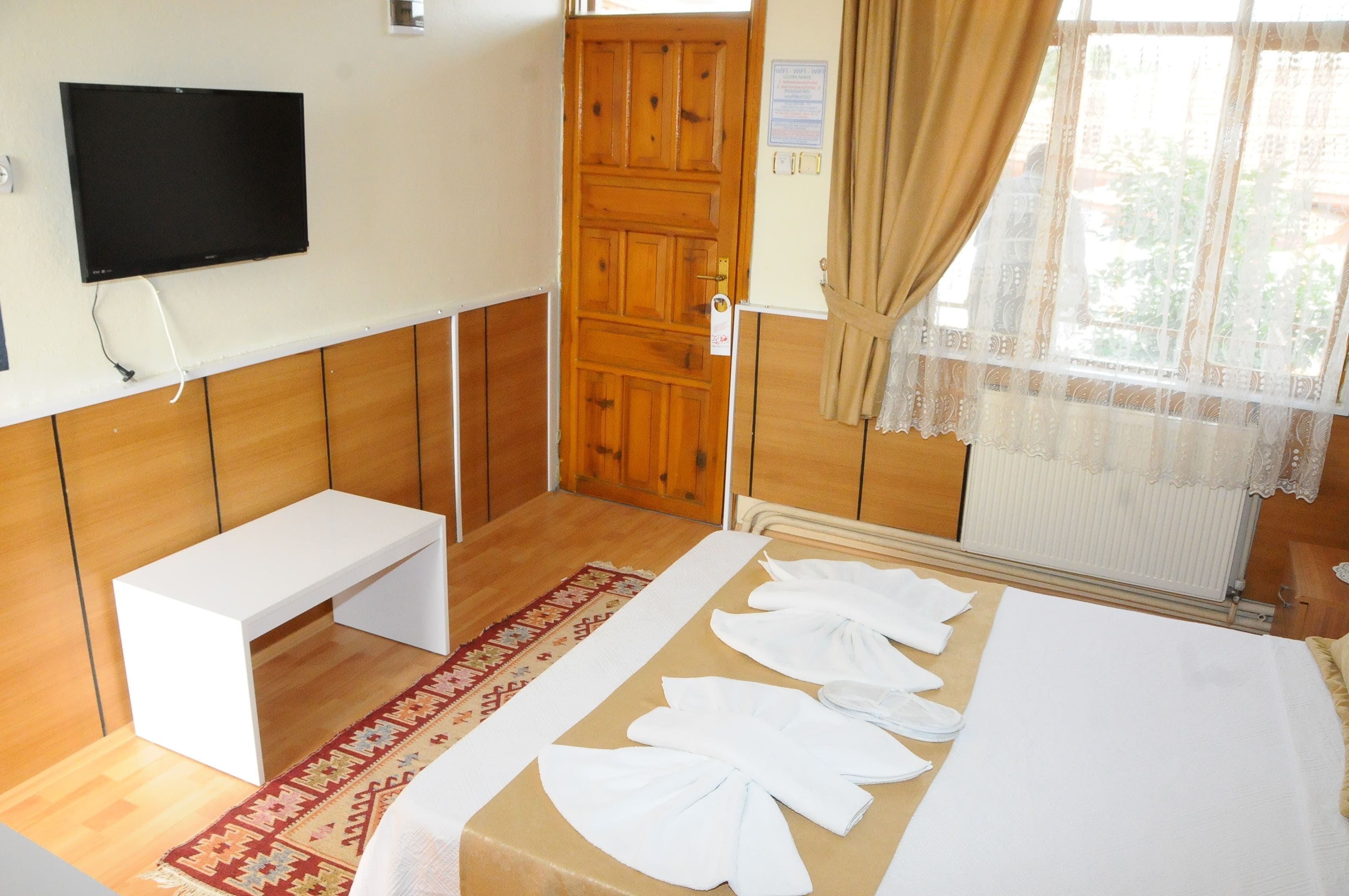 A hotel room with a wooden floor, a white bench, a bed with towels, a TV on the wall, and a window with curtains.