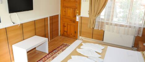 A hotel room with a wooden floor, a white bench, a bed with towels, a TV on the wall, and a window with curtains.