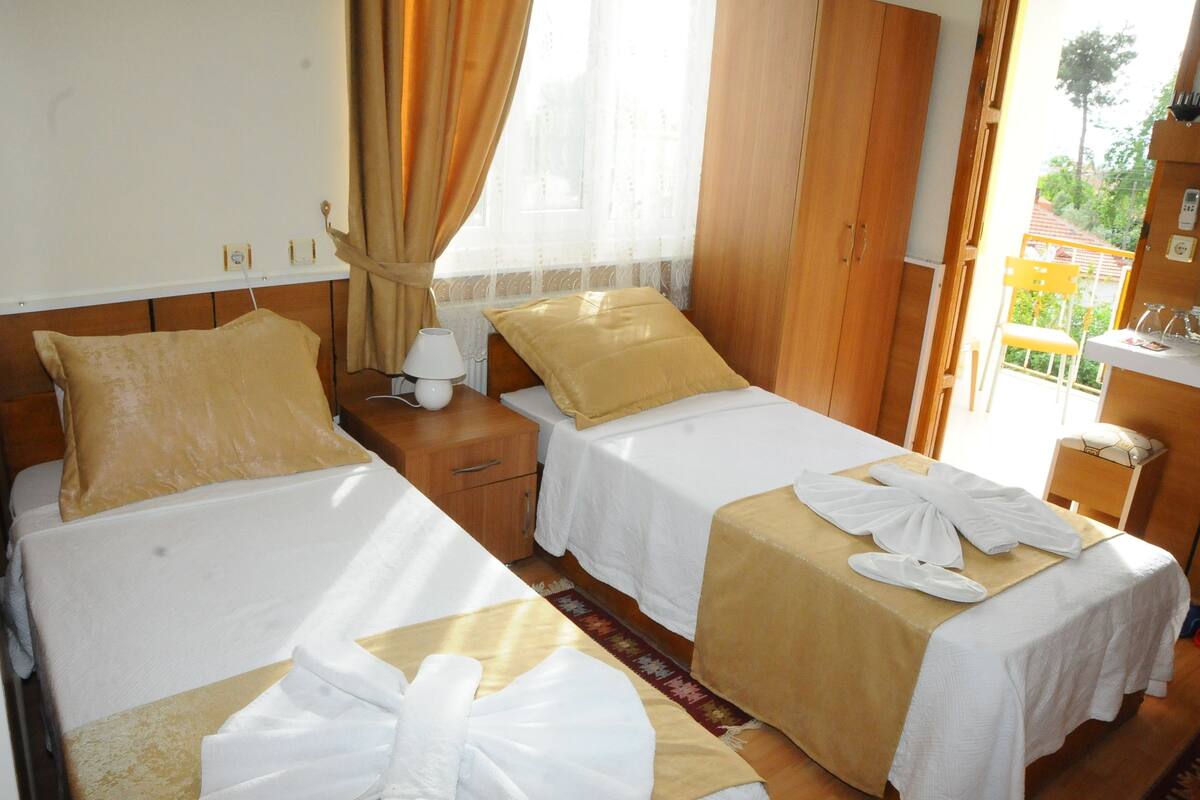 A hotel room with two beds, each with a white bedspread and a beige pillow. There is a wooden nightstand with a lamp and a small table with a glass of water and a plate of fruit. The room has wooden walls and a window with curtains.