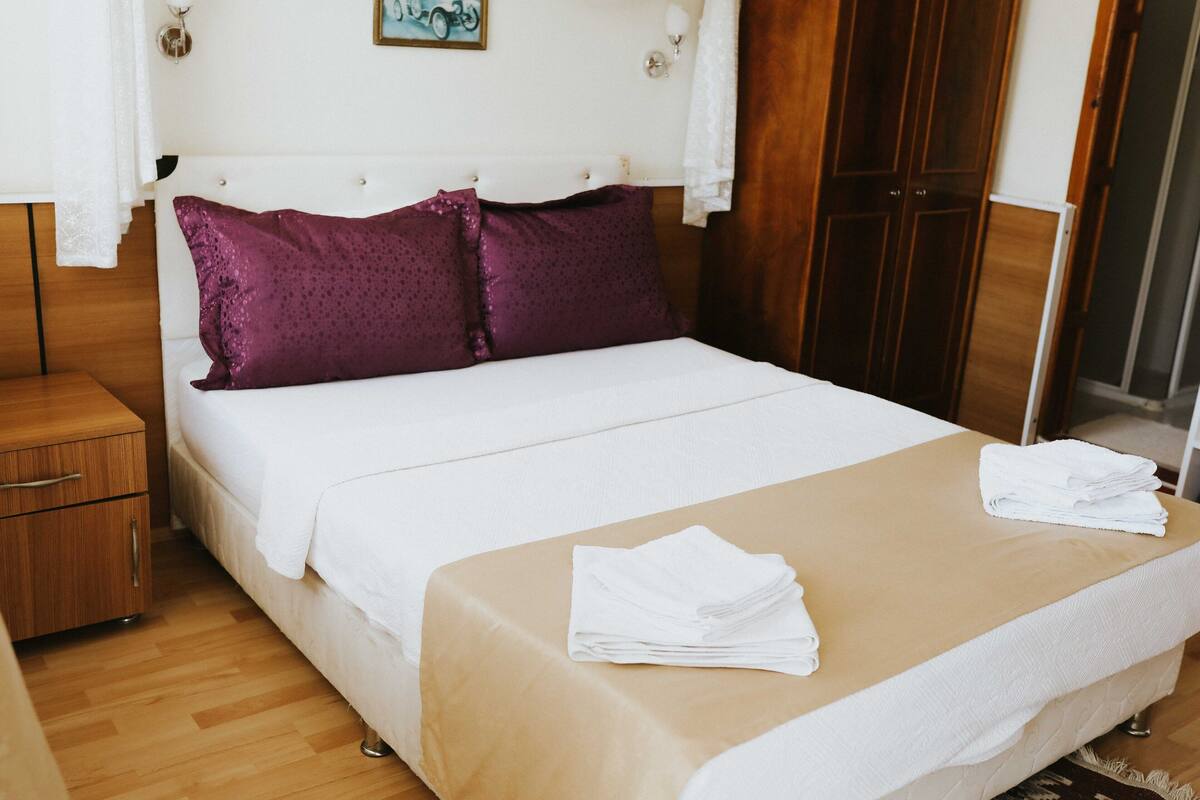 A neatly made bed with a white and beige bedspread, two purple pillows, and a stack of white towels on the bed.