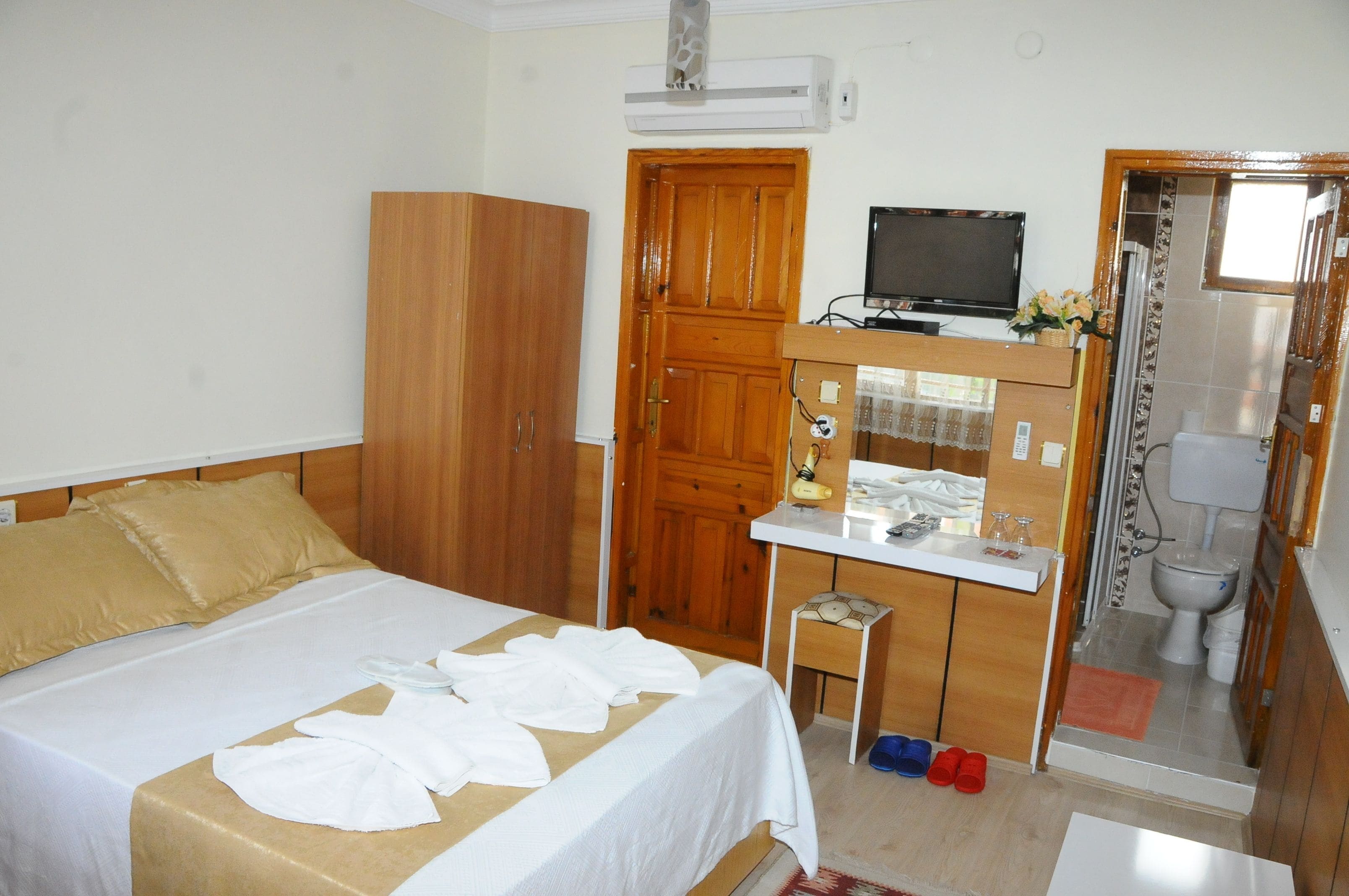 A hotel room with a bed, wooden wardrobe, a desk with a television, a small table with toiletries, and a bathroom visible through an open door.