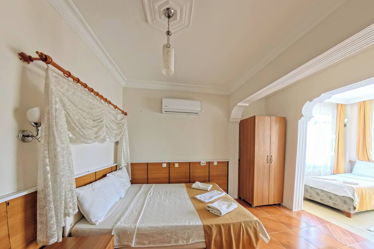 A hotel room with a bed, a wooden wardrobe, a nightstand, a bedside table, and an air conditioning unit.