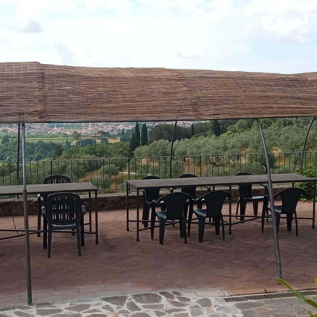 Une terrasse extérieure avec des tables et des chaises, une pergola offrant de l’ombre et une vue sur un paysage verdoyant, avec des arbres et des bâtiments visibles au loin.