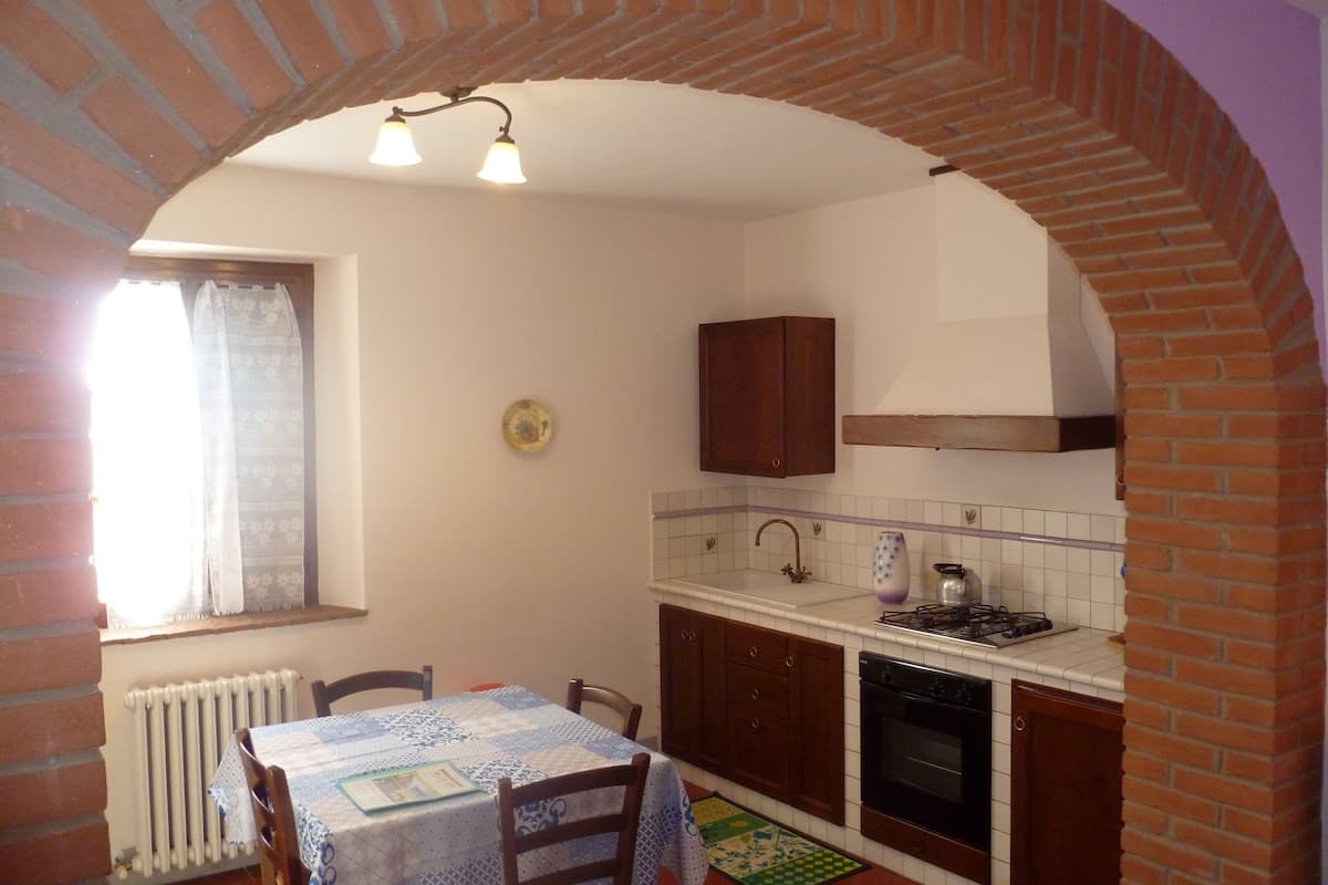Una cocina con arco de ladrillo, una ventana con cortinas, una mesa con sillas, un radiador y una estufa.
