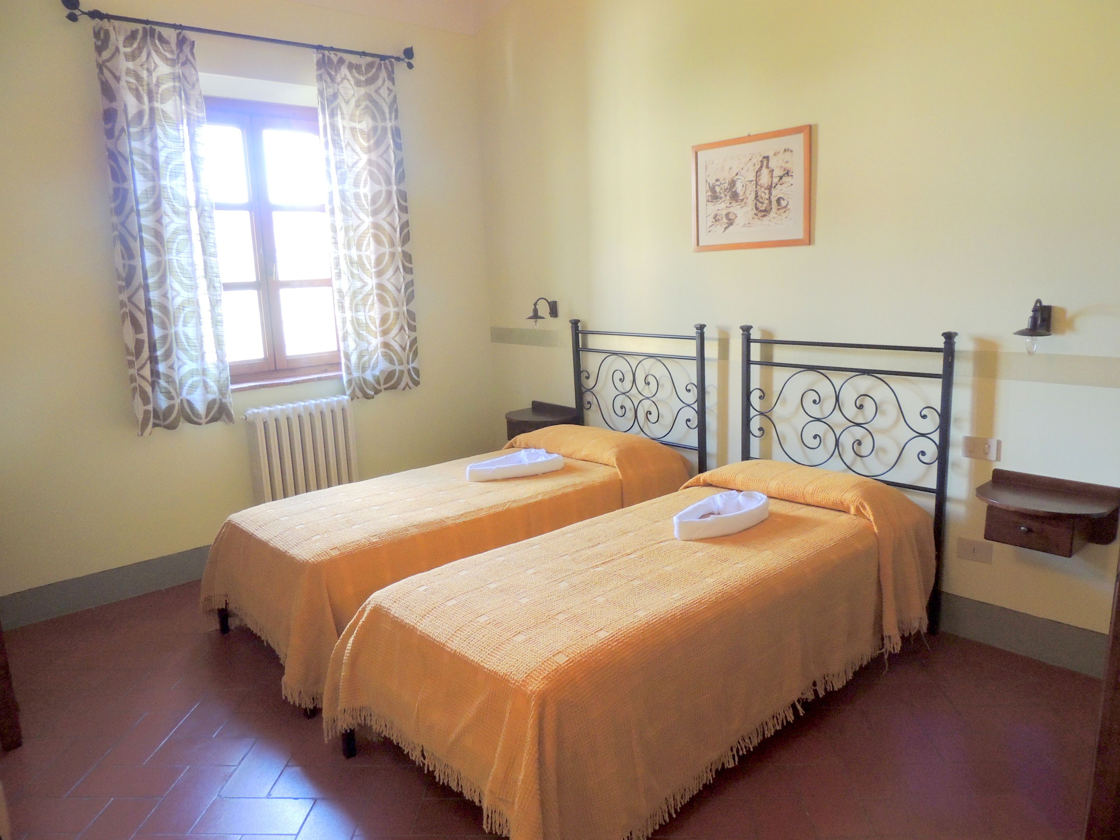 A room with two beds, a window with curtains, a radiator, a framed picture on the wall, and a bedside table with a lamp.