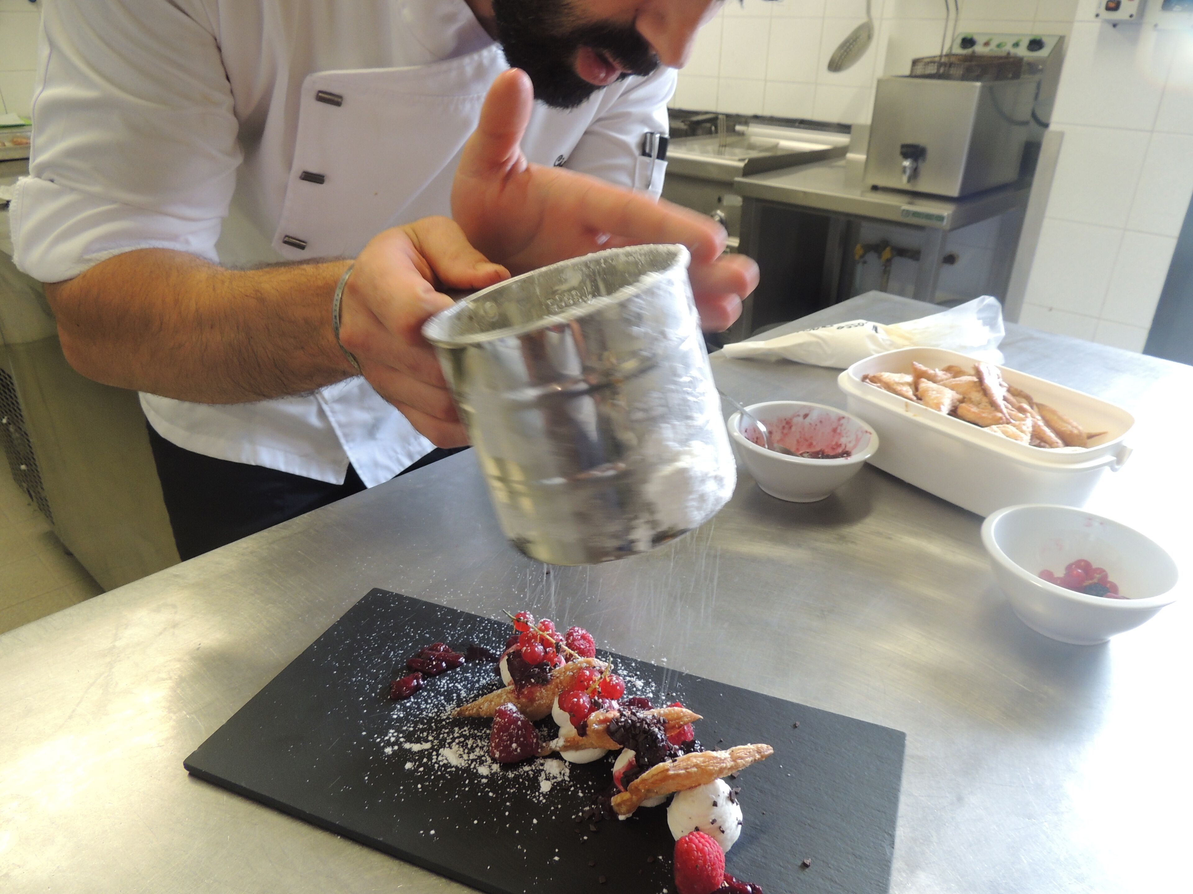 A chef preparing a dessert on a black slate plate, with a bowl of berries and a container of powdered sugar nearby.