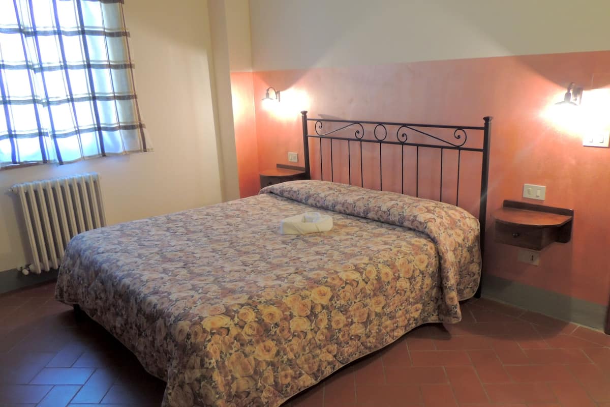 Un dormitorio con colcha estampada, un radiador, una ventana con persianas y un estante fijado a la pared.