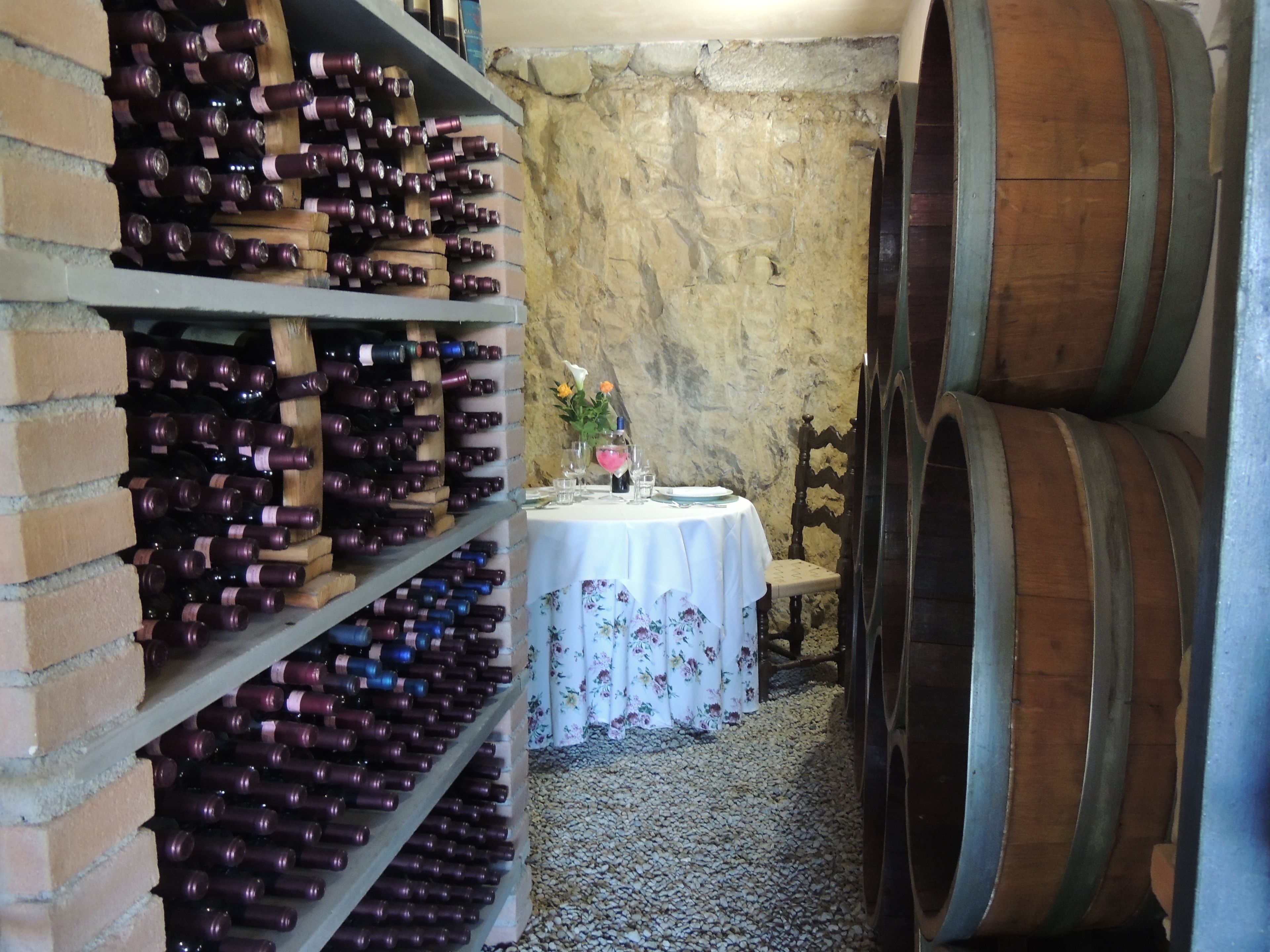 Une cave à vin avec des étagères en bois où sont rangées les bouteilles, une table recouverte d’une nappe à motifs floraux et une chaise.