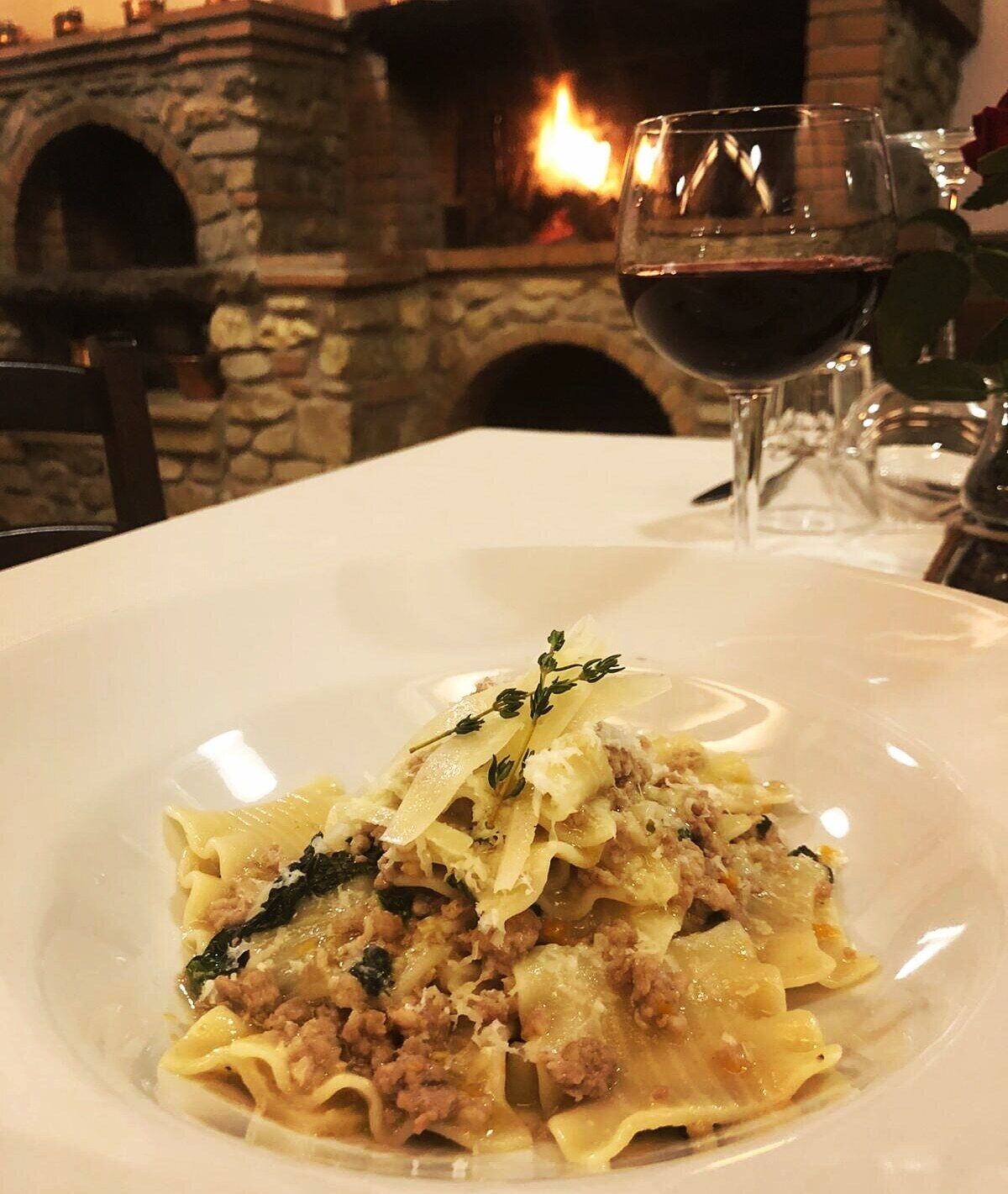 A plate of pasta with a garnish, a glass of red wine, and a stone fireplace in the background.