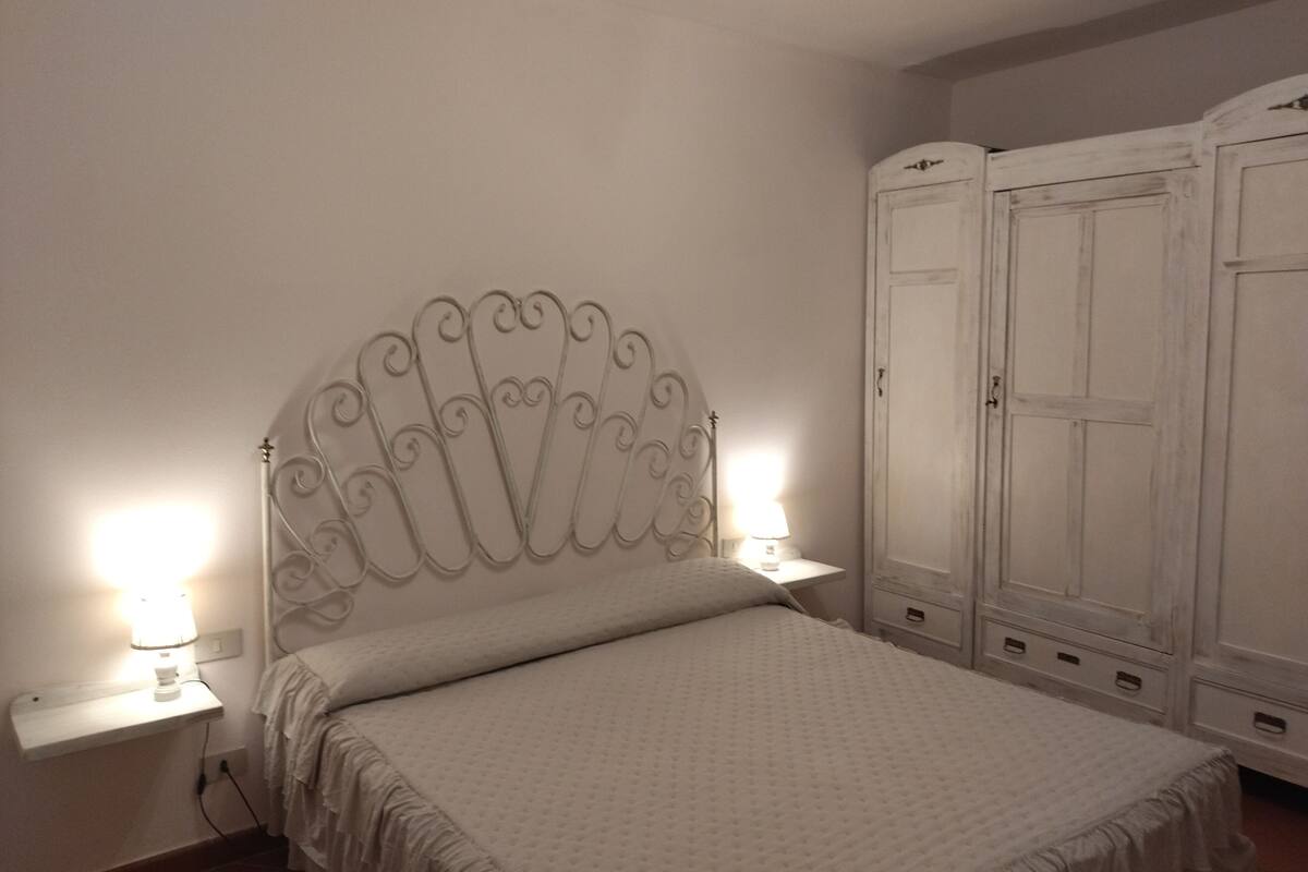 Un dormitorio con una cama blanca, un cabecero decorativo, dos lámparas de noche y un armario.