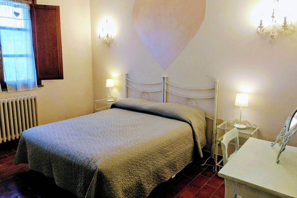 Un dormitorio con cama, dos mesitas de noche con lámparas, un radiador, una ventana con cortinas y una pared con un diseño de corazón.