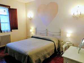 Une chambre avec un lit, deux tables de chevet avec des lampes, un radiateur, une fenêtre avec des rideaux et un mur décoré d’un motif en forme de cœur.