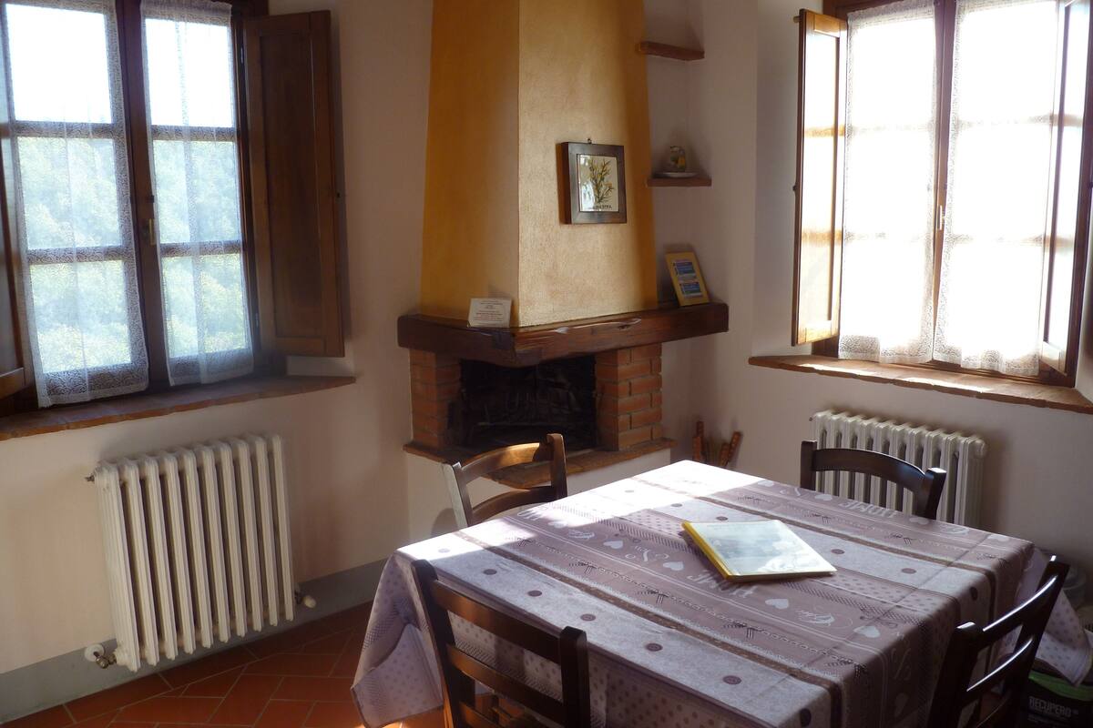 Un comedor con una mesa preparada para dos personas, chimenea y ventana con cortinas de encaje.