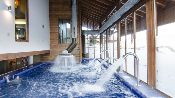 A hot tub with multiple water jets, wooden beams, and a view of the outside through large windows.