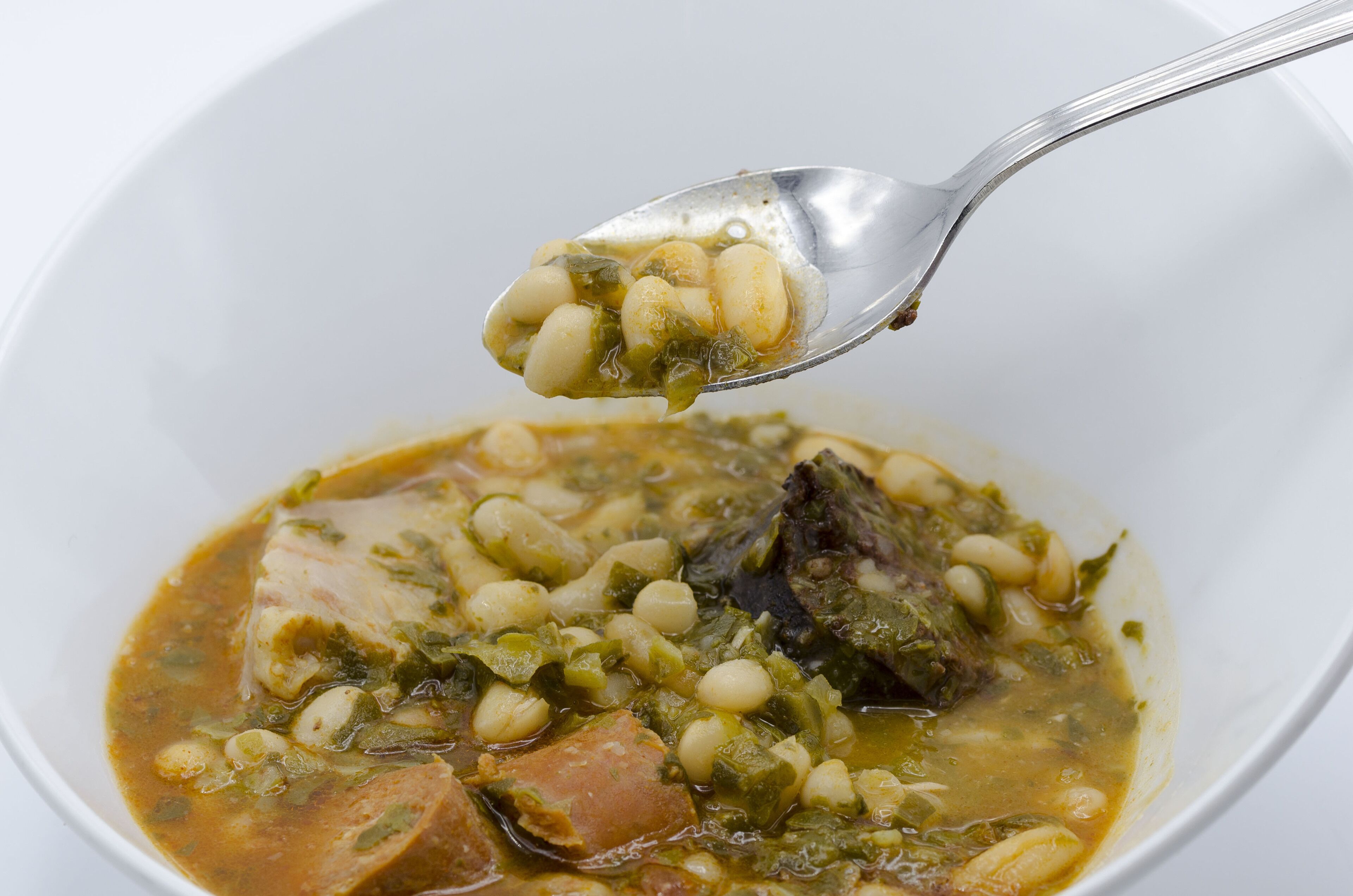 Un tazón de sopa con ingredientes visibles como frijoles, verduras de hoja verde y trozos de carne.