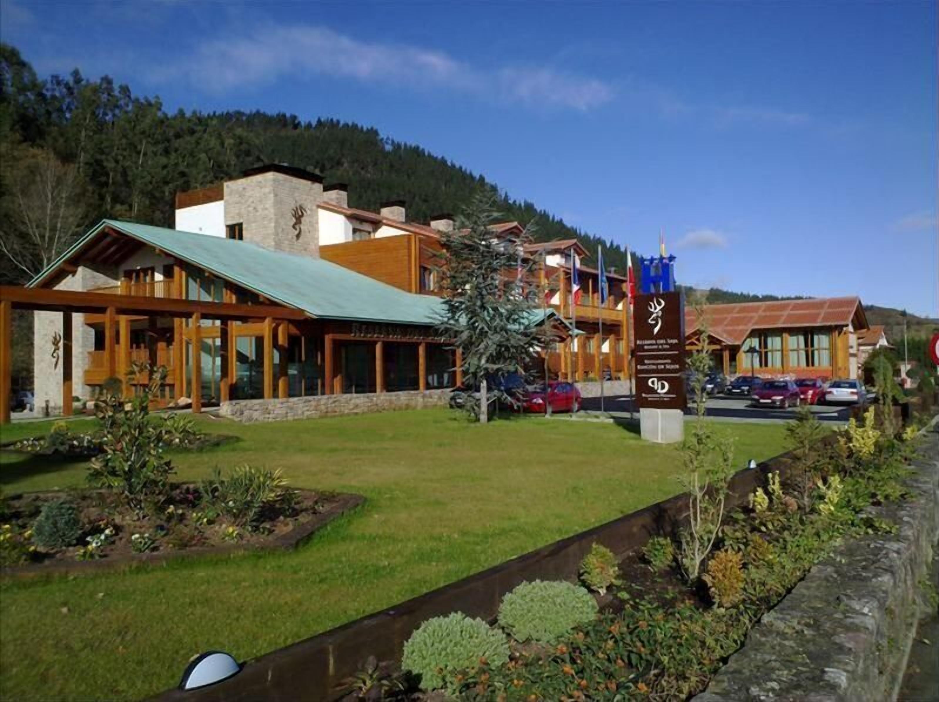Un hotel moderno con un amplio frente de vidrio, techo verde y base de piedra. Hay varias banderas y un cartel informativo frente al edificio.