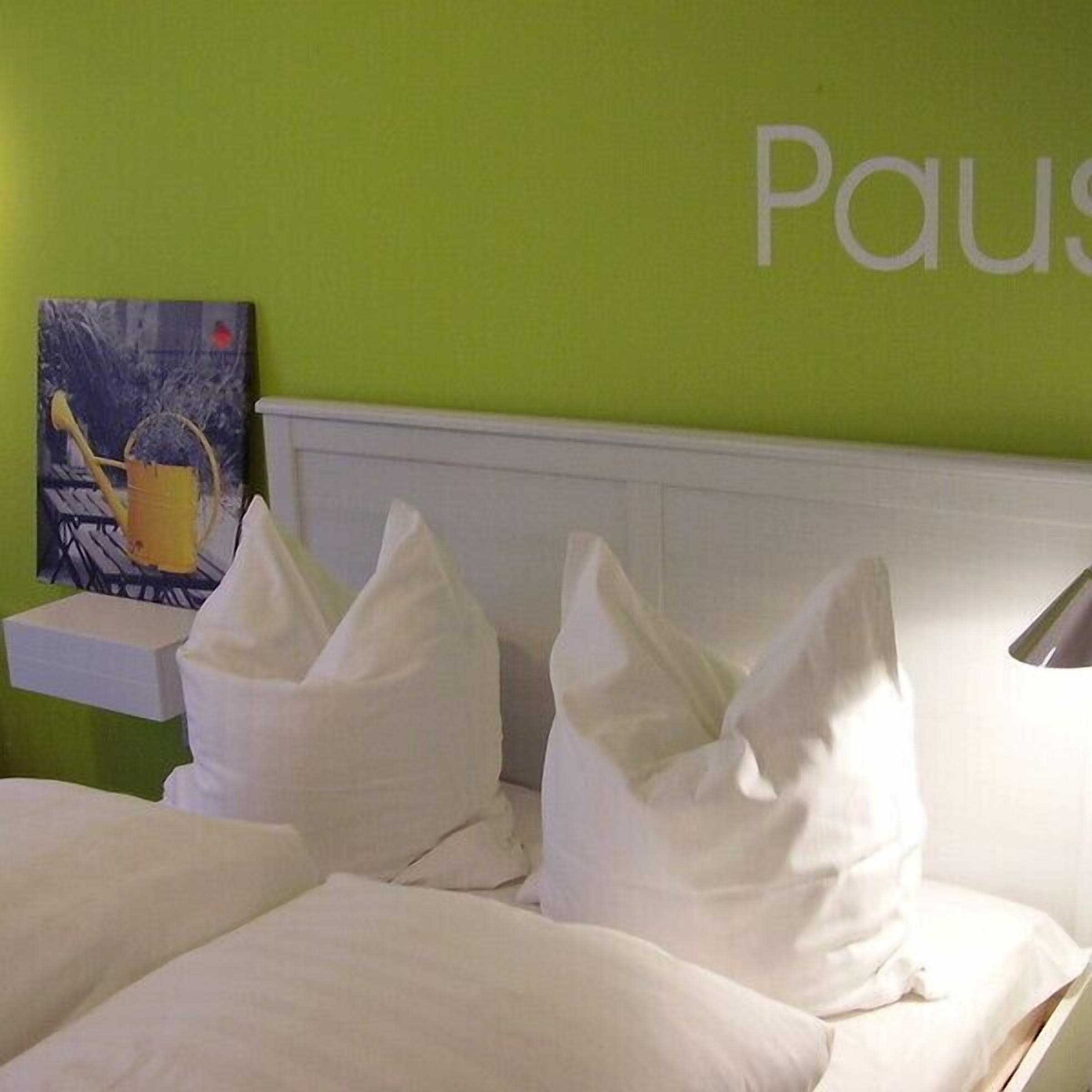 A hotel room with a green wall, a white headboard, and a painting of a watering can.