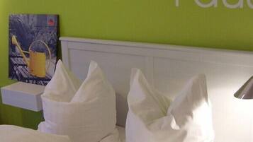 A hotel room with a green wall, a white headboard, and a painting of a watering can.