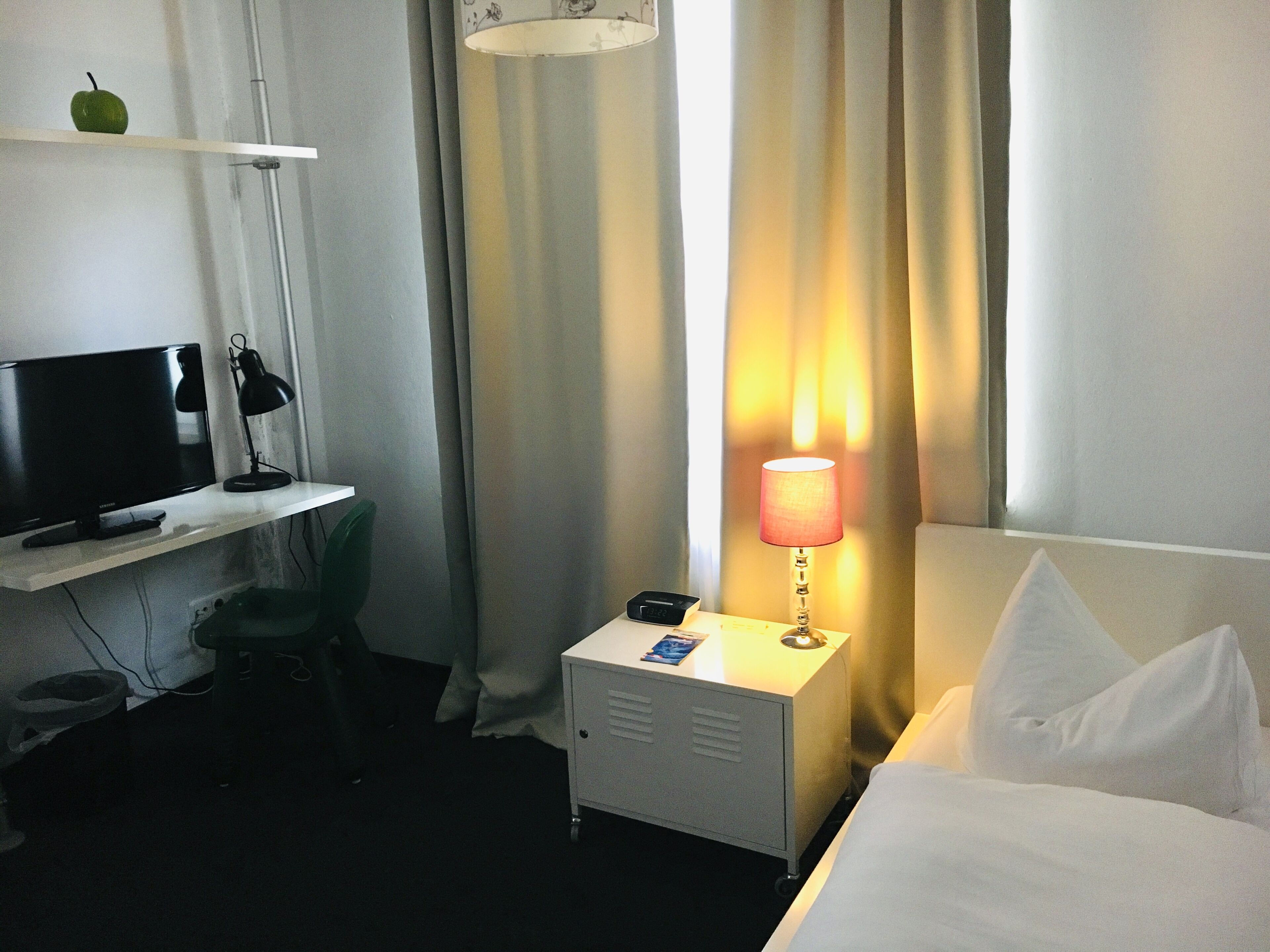 Een hotelkamer met een bed, een bureau met televisie en lamp, een klein kastje en een raam met gordijnen.