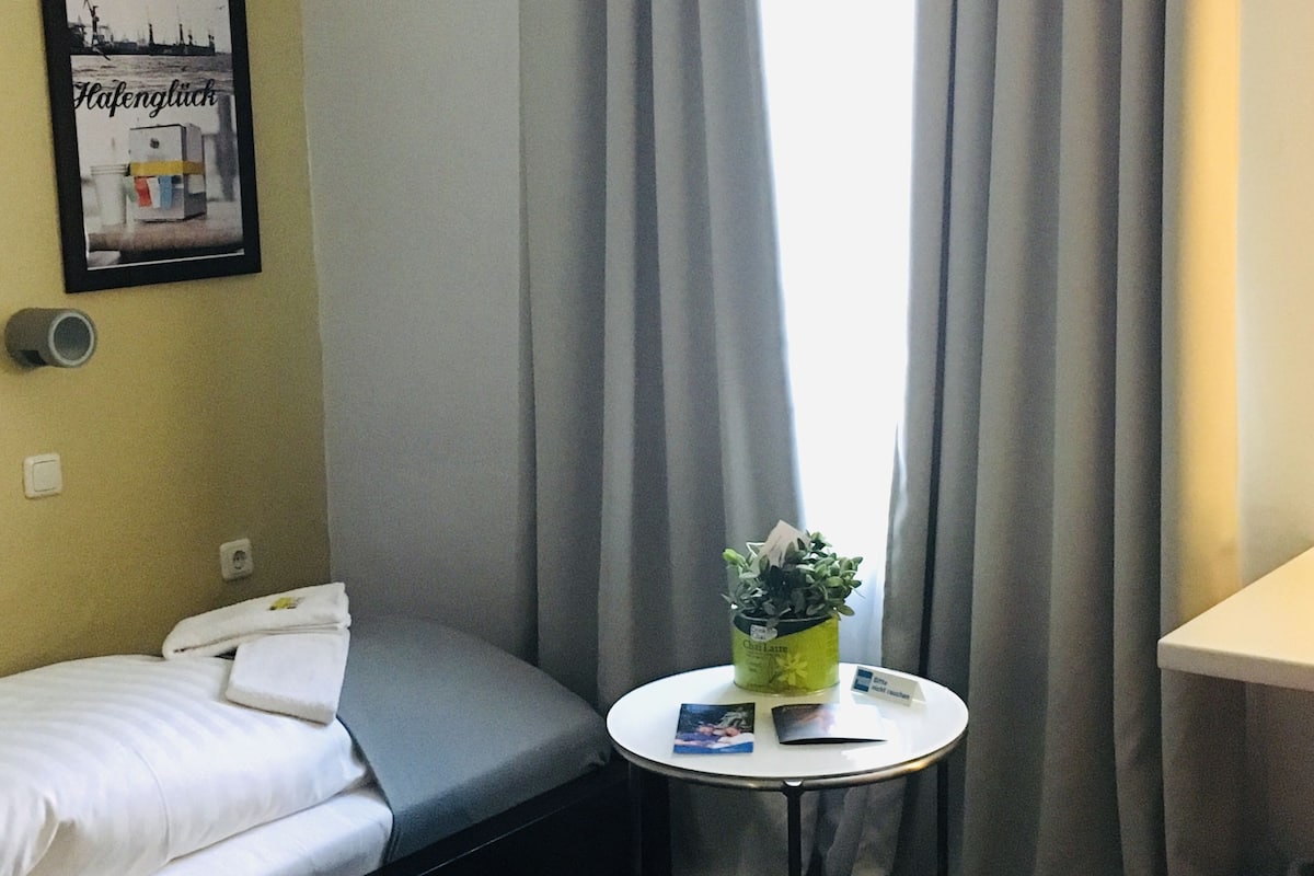 Hotellihuone, jossa on sänky, pieni pöytä, jossa on kasvi, seinällä oleva kehystetty kuva ja ikkuna, jossa on verhot.