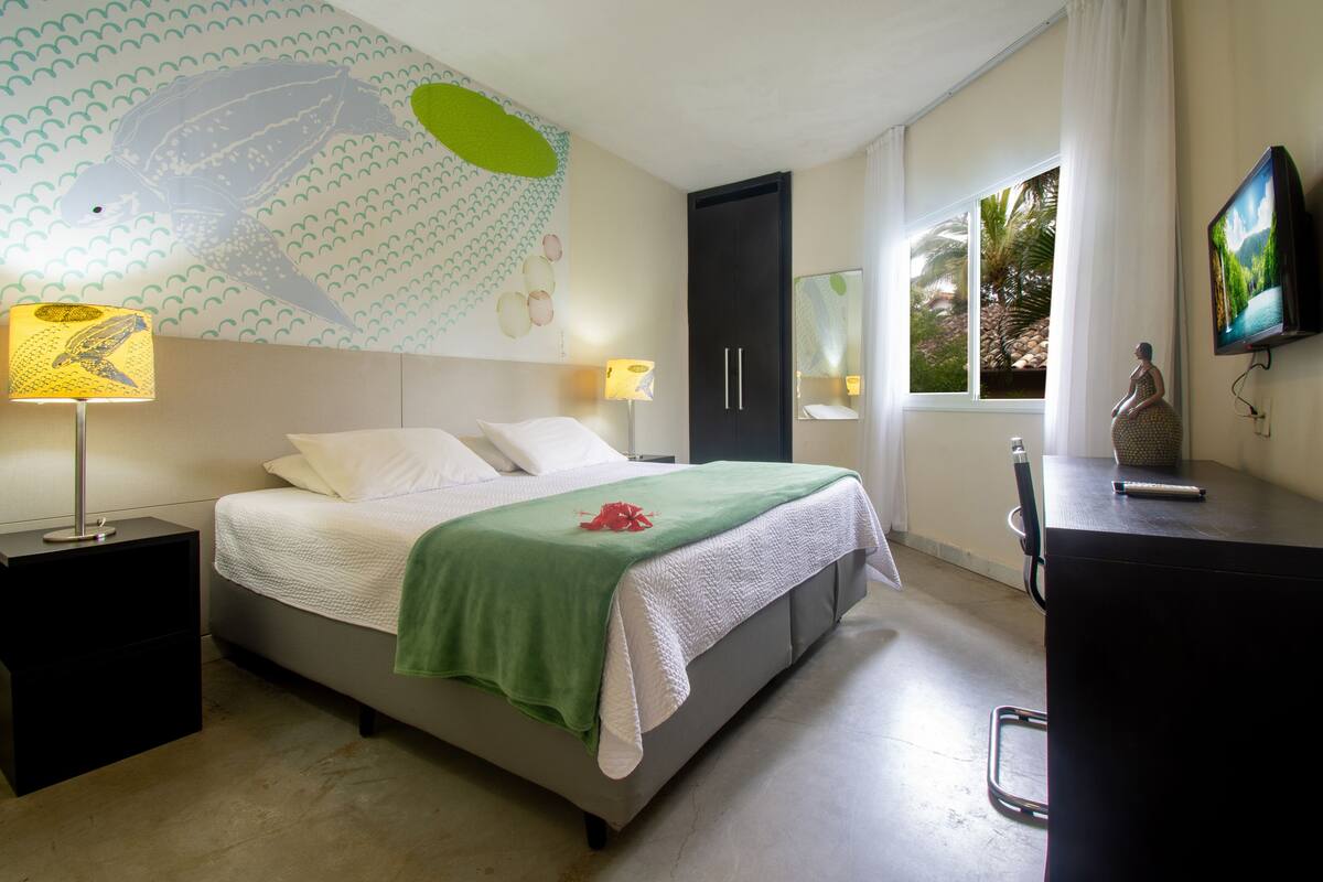 A hotel room with a bed, nightstands, a desk, a TV, and a large window with a view of palm trees.