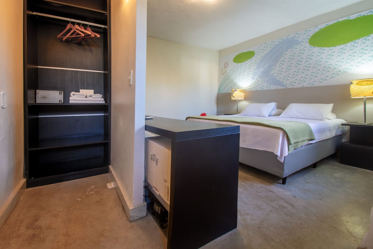 A hotel room with a bed, a desk, a wardrobe, and a wall with a green abstract design.