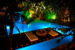 Uma área de piscina interior com pouca luz, folhagem tropical e uma mesa posta com um livro e um objeto decorativo.