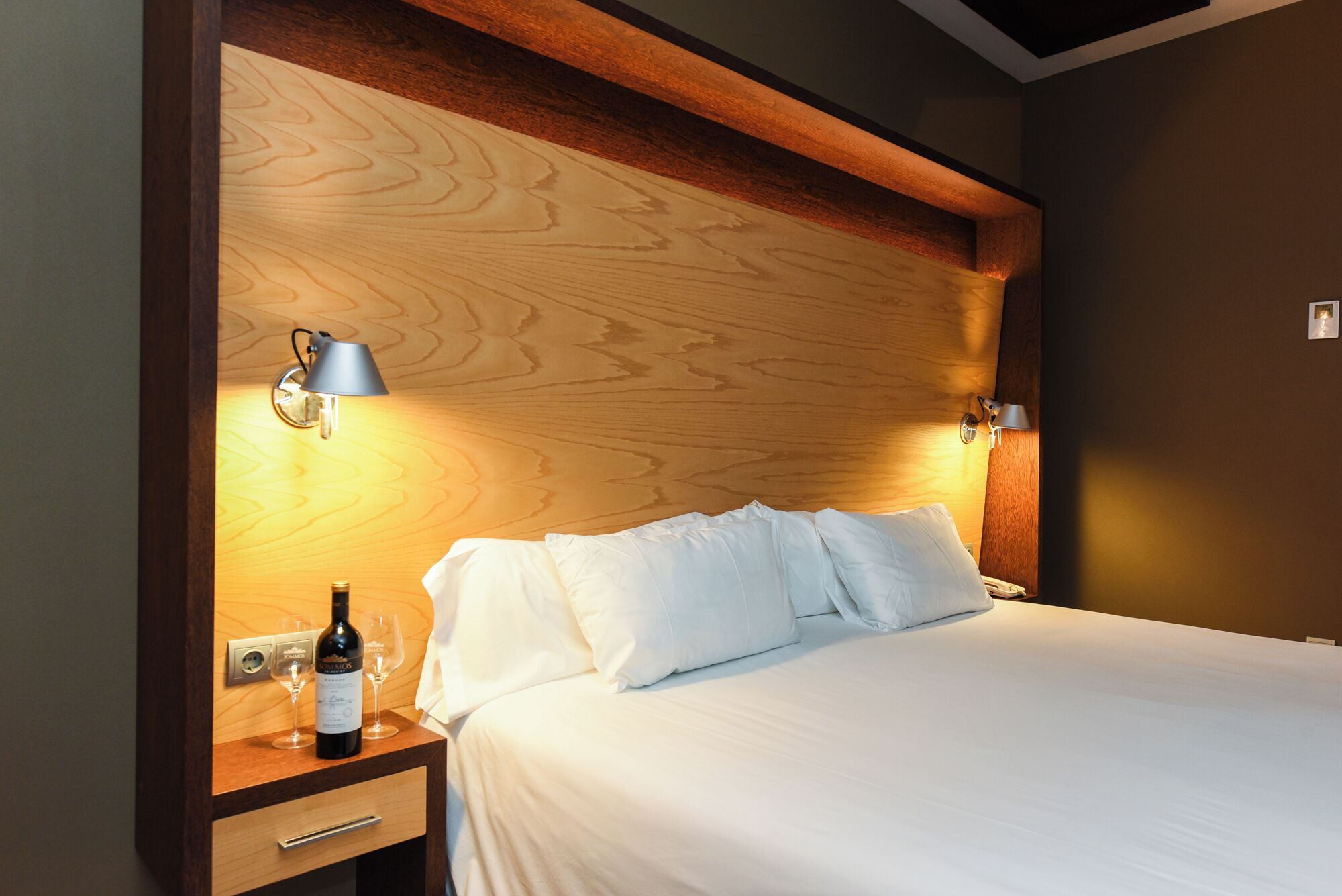 A hotel room with a wooden headboard, two bedside lamps, a bed with white linens, a nightstand with a wine bottle and glasses, and a drawer.