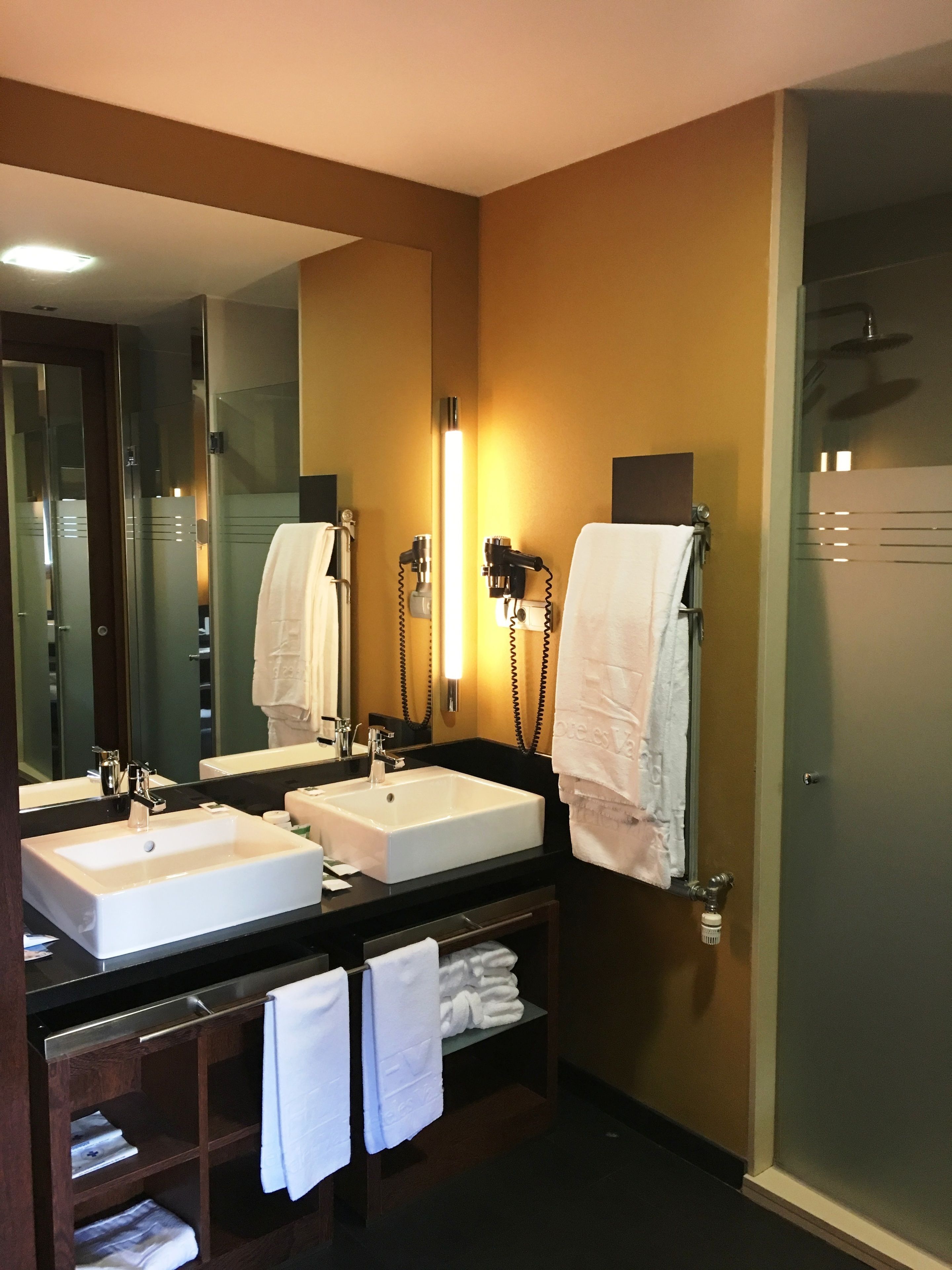 A hotel bathroom with two sinks, a large mirror, and a hairdryer.
