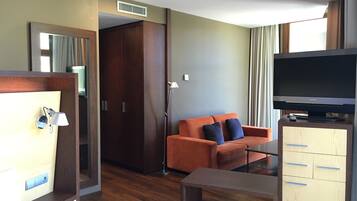 A modern hotel room with a wooden wardrobe, a sofa with cushions, a small table, a TV on a stand, and a large window with curtains.